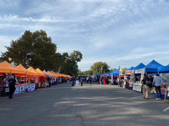 2026年10月10日下午，Queens Night Market 的道路两侧排列橙色和蓝色摊棚，游客在中间通行