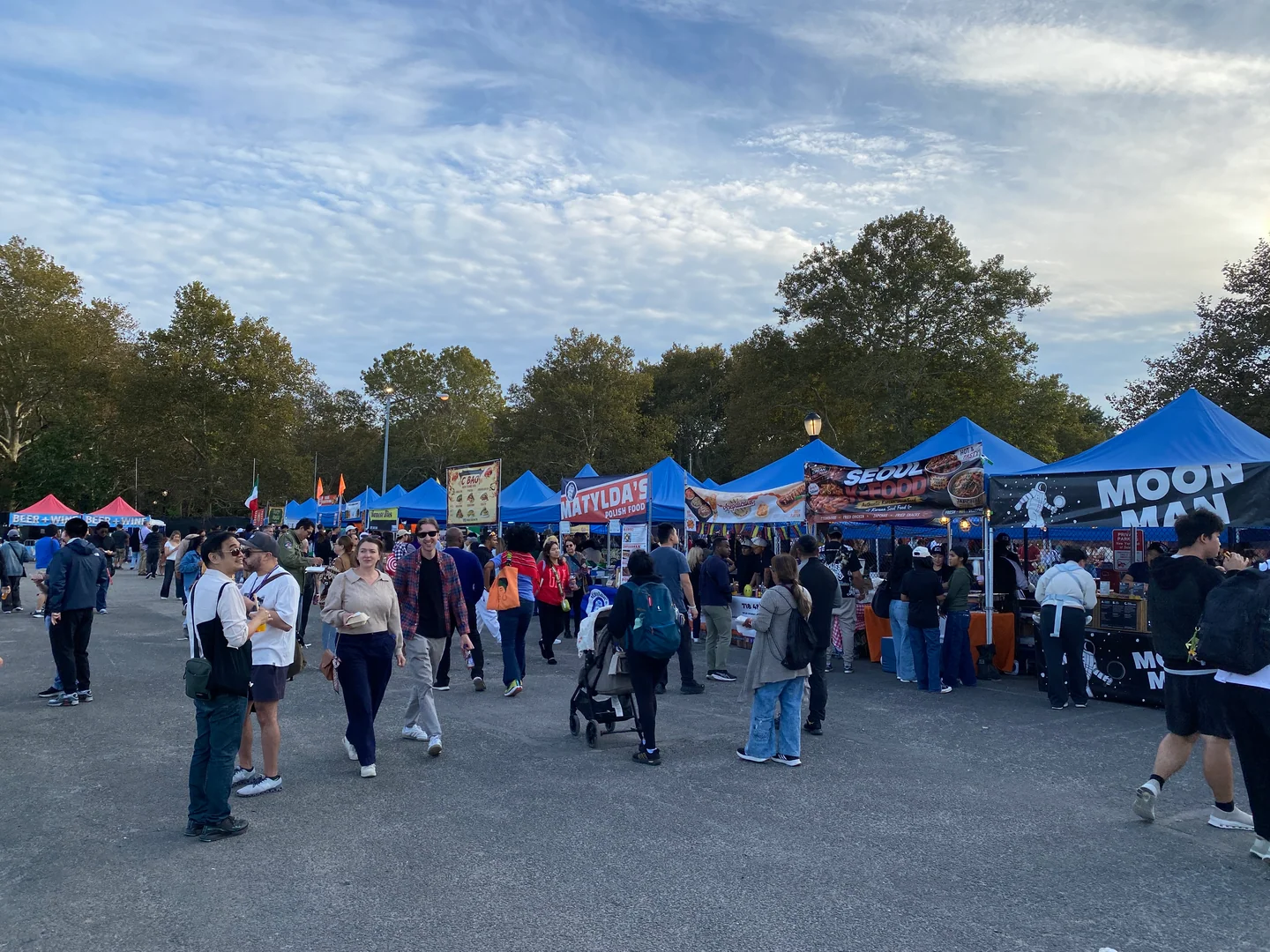 2026年10月10日下午，游客在 Queens Night Market 的蓝色摊棚前走动，前景有多名行人