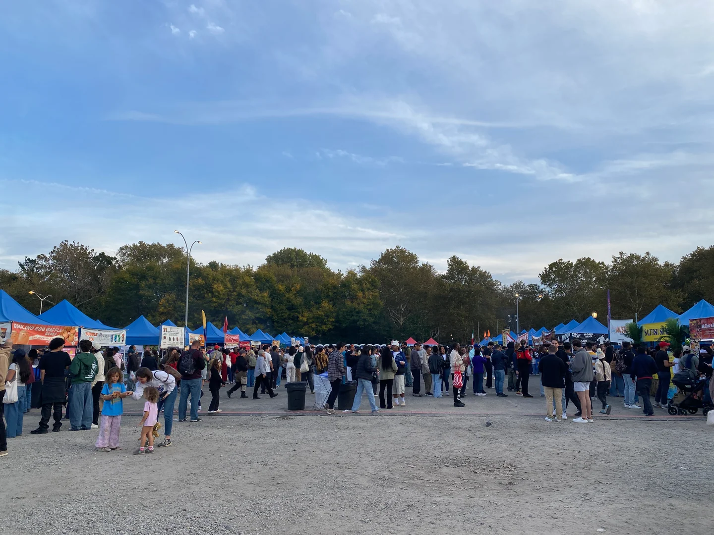 2026年10月10日傍晚，Queens Night Market 的摊棚之间聚集游客，远处可见两排蓝色棚顶